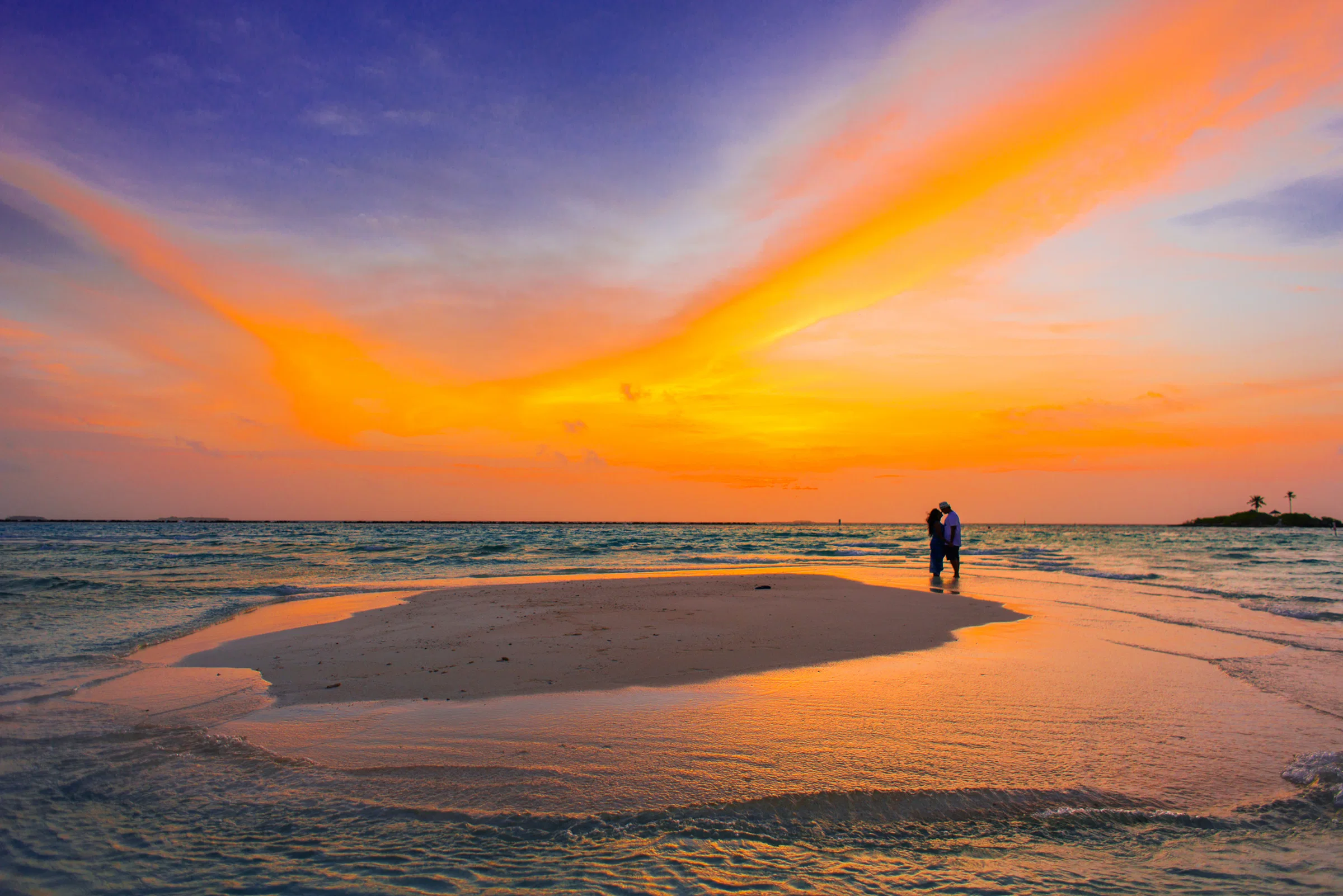 An orange sunset over a sandbank and shallow water, two small figures in silhouette