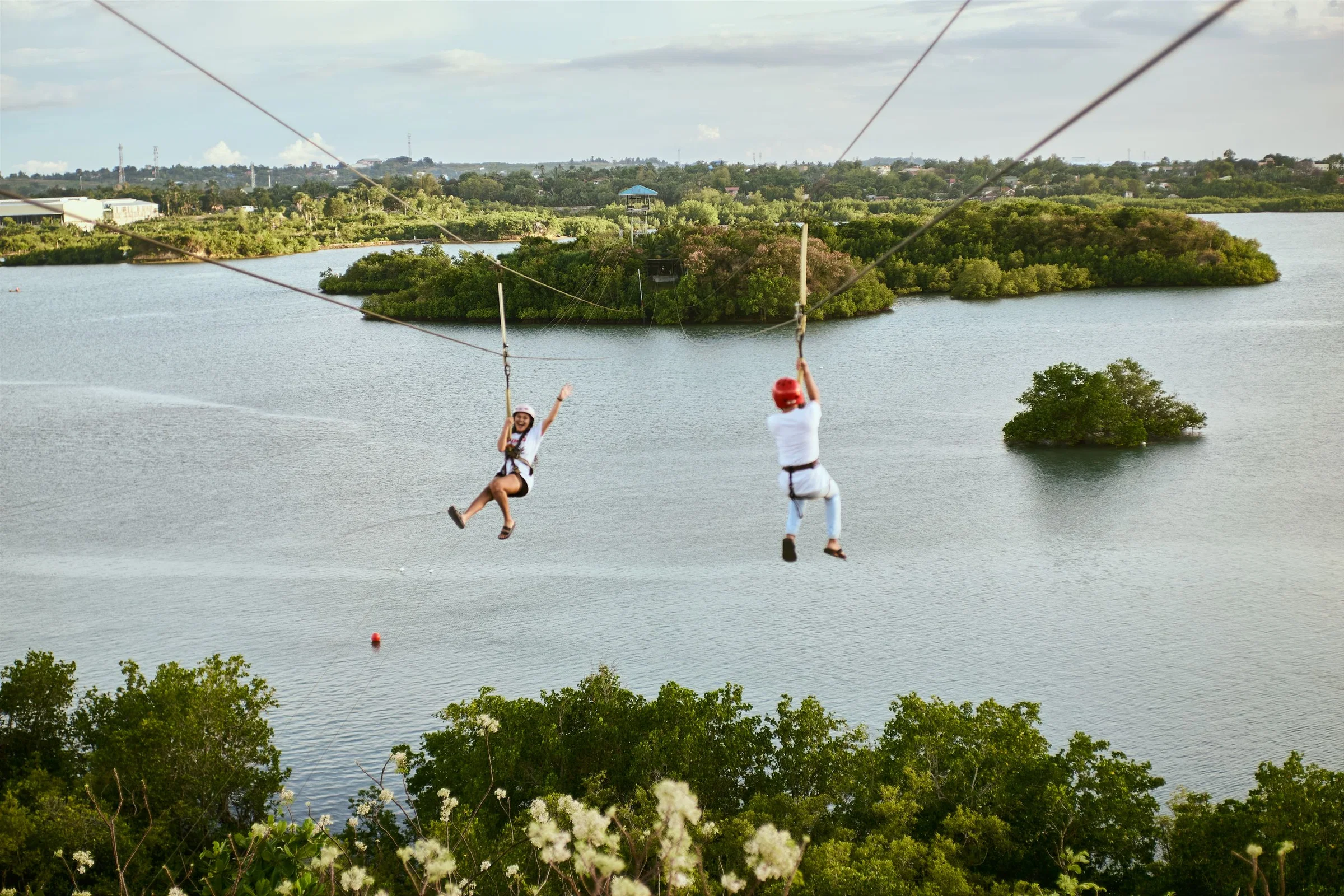 Two riders on a zipline over water and green islets, seen from a distance
