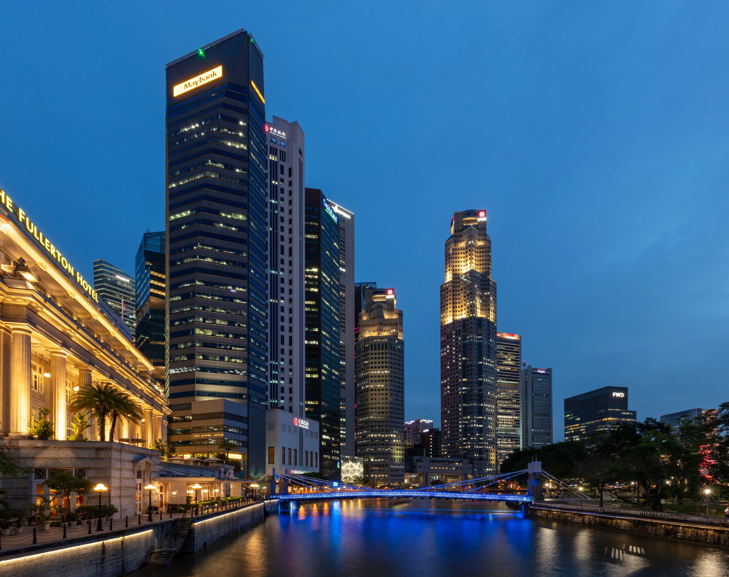 The illuminated Fullerton Hotel beside the Singapore River at blue hour, with the towers of the financial district behind