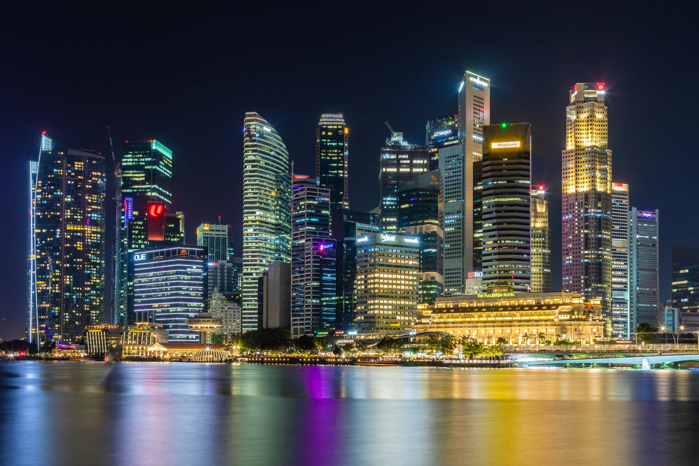 The towers of Singapore's Central Business District lit at night and reflected in Marina Bay, with the Fullerton Hotel at the waterline