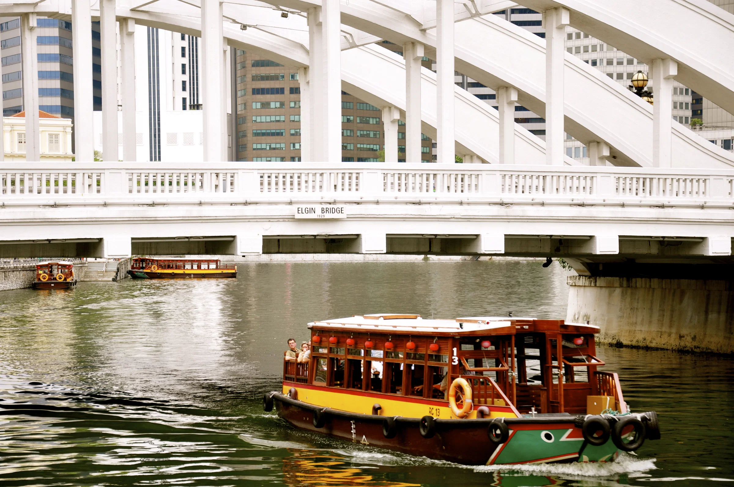 A traditional wooden bumboat passing beneath the white arches of Elgin Bridge on the Singapore River