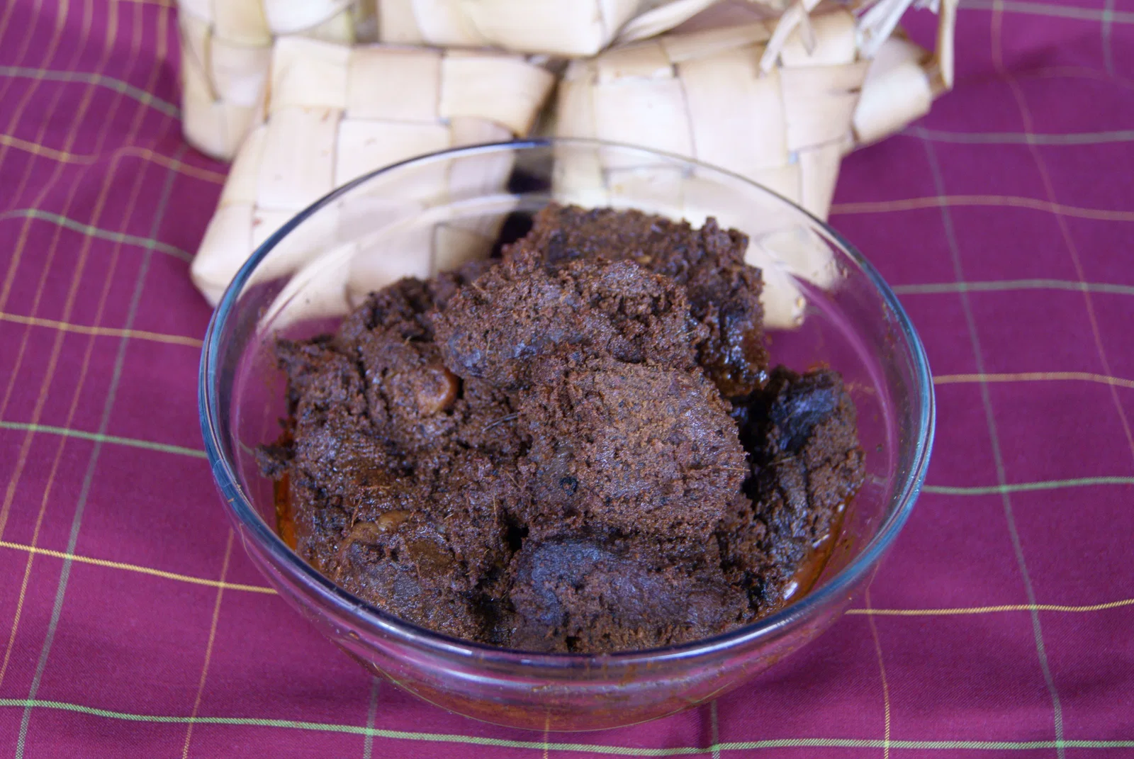 A glass bowl of dark beef rendang beside woven ketupat rice parcels