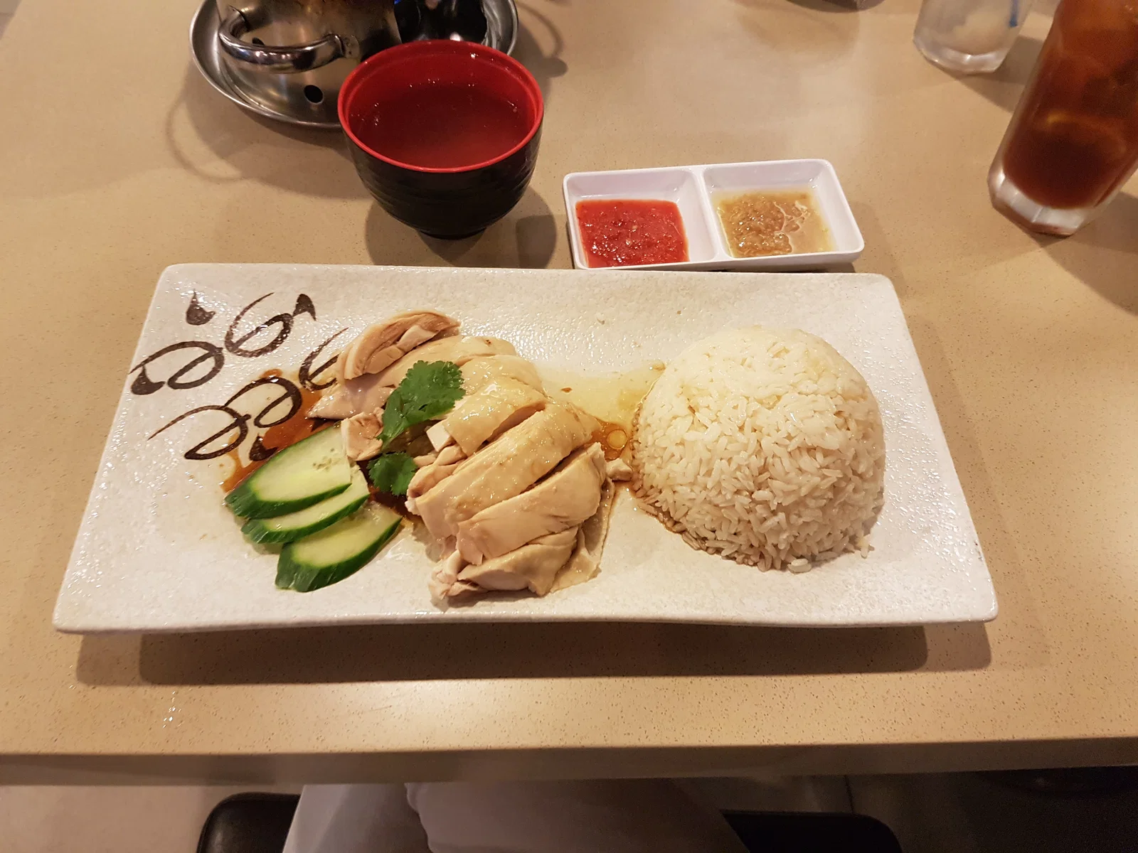 A plate of Hainanese chicken rice with sliced poached chicken, cucumber, rice and dipping sauces