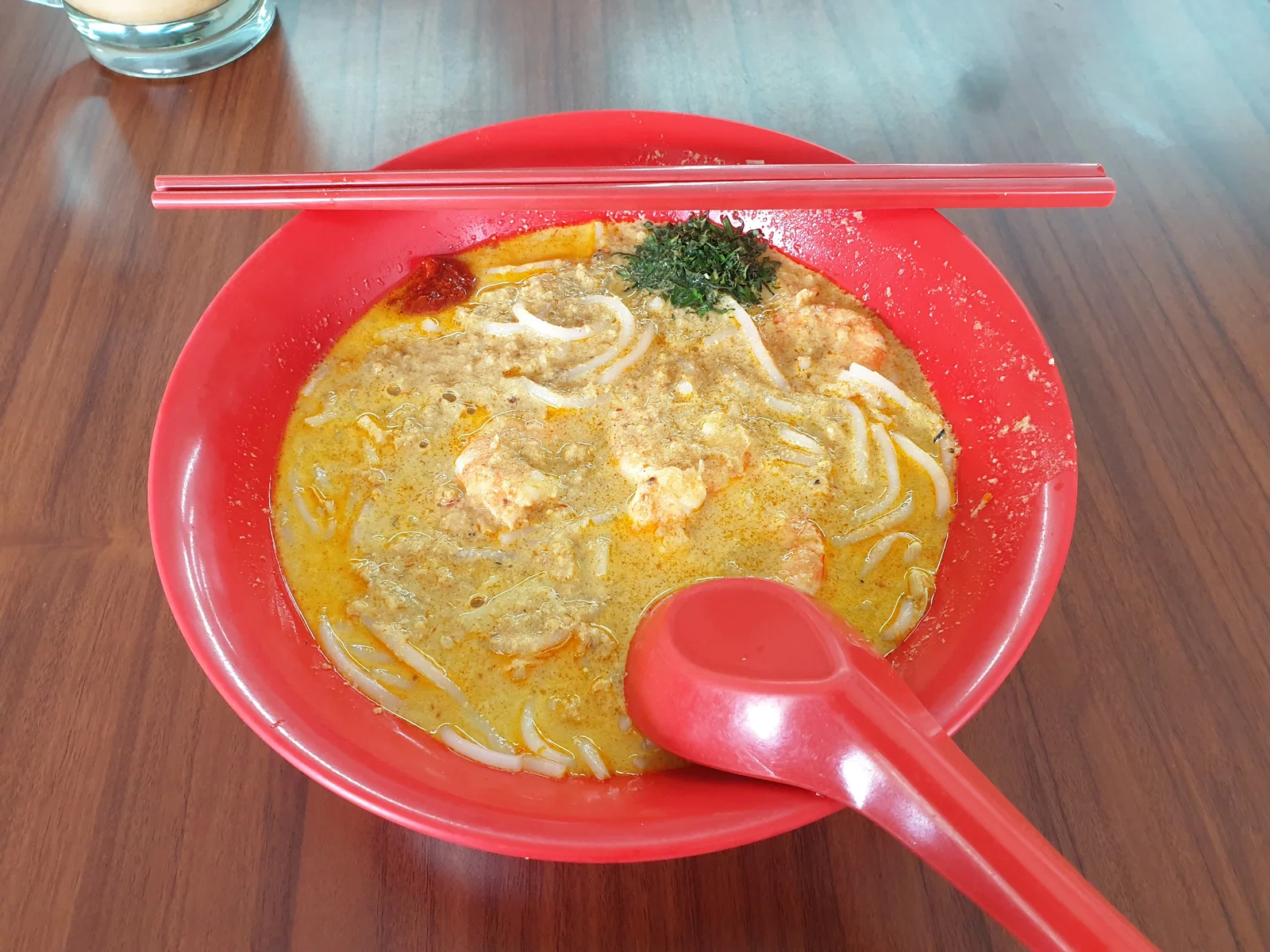 A red bowl of Katong laksa with coconut broth and short noodles, with a spoon and chopsticks, in Singapore