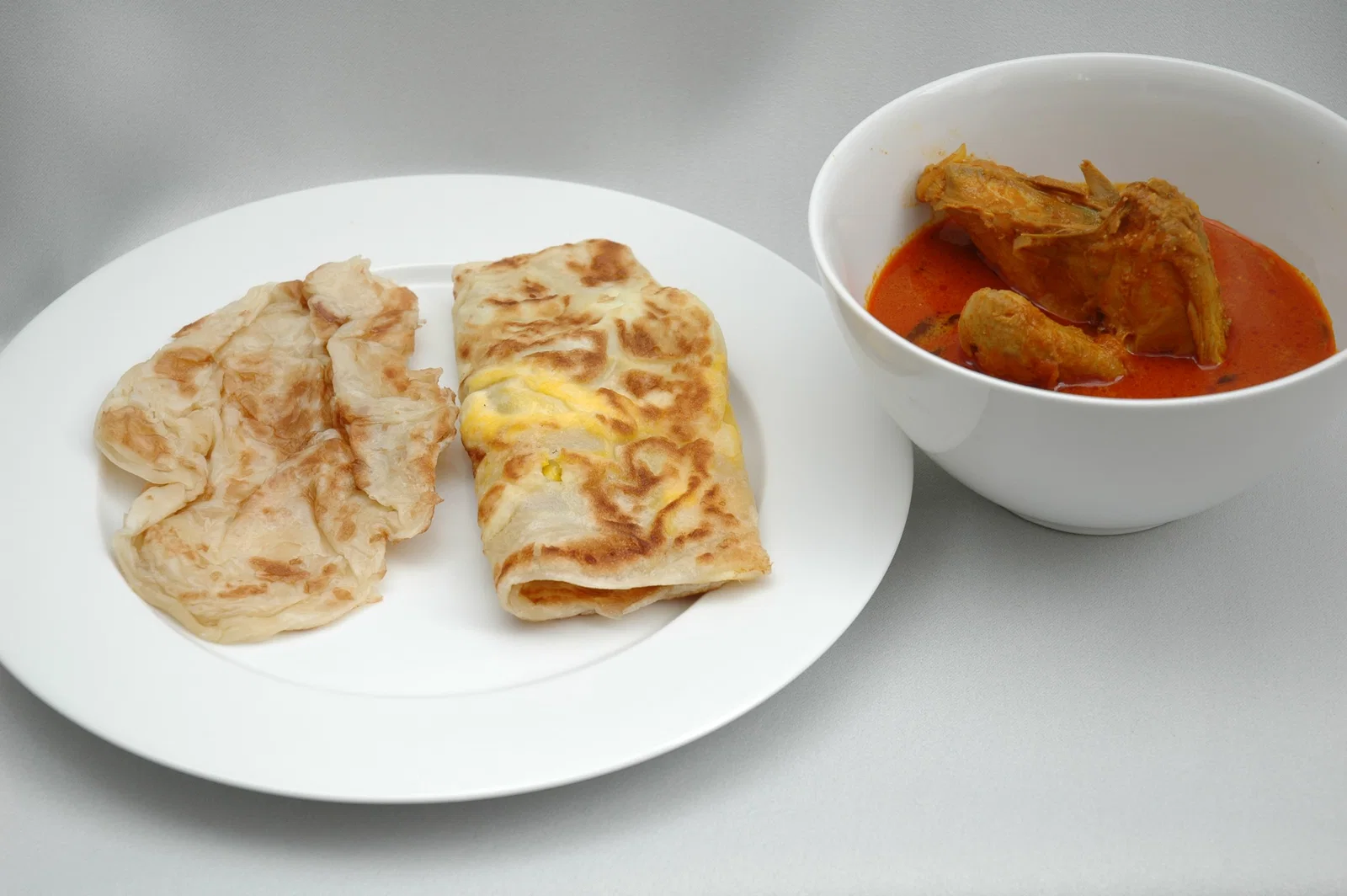 Plain and egg roti prata on a plate beside a bowl of chicken curry