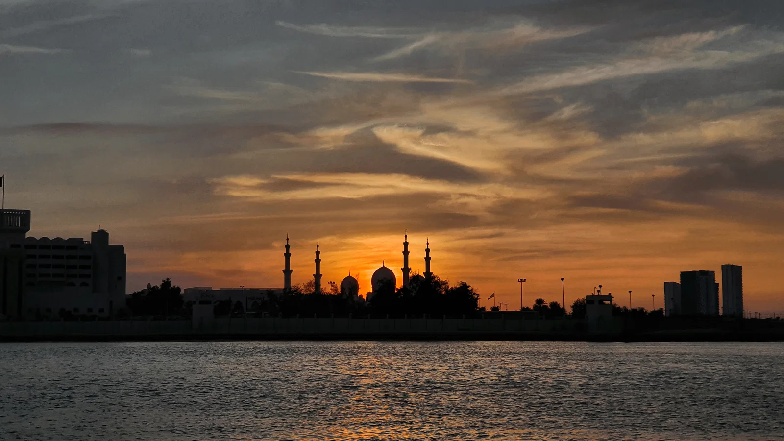 The domes and minarets of the Sheikh Zayed Grand Mosque in silhouette at sunset across the canal